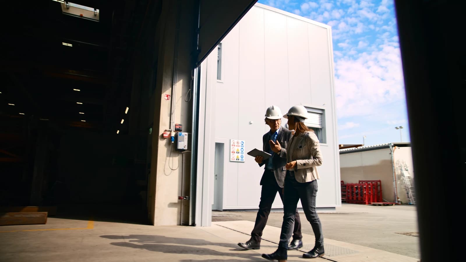 Two people in hard hats walking out through a warehouse dock door, one carrying a tablet